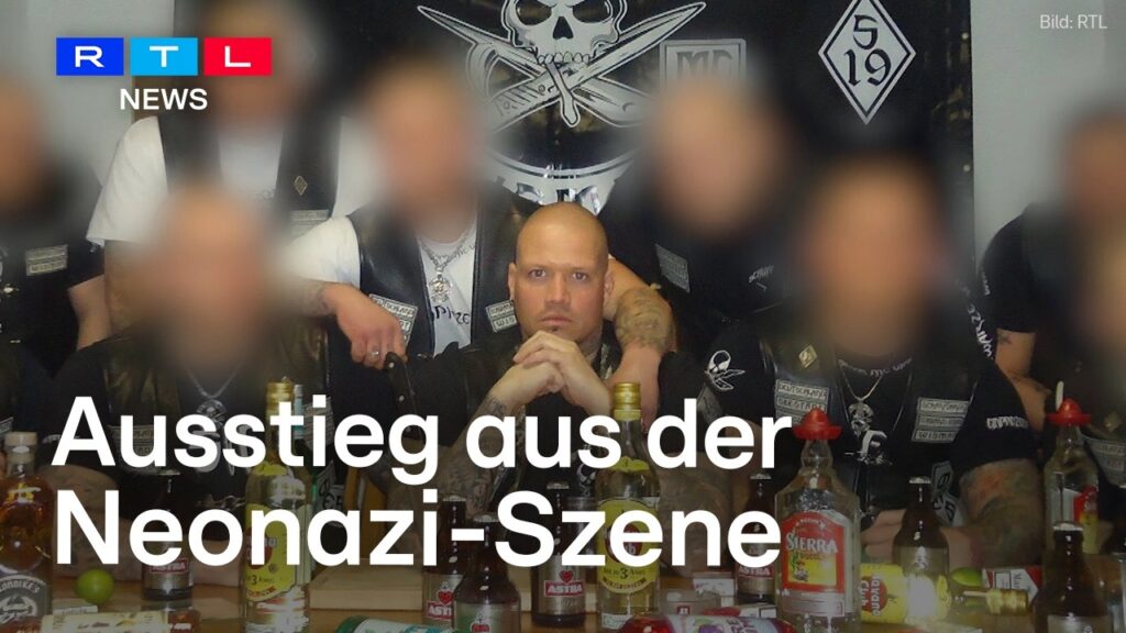 Ein Mann mit Glatze, möglicherweise Philip Schlaffer, sitzt an einem Tisch mit Flaschen und Gläsern, umgeben von anderen Männern, deren Gesichter unscharf sind. Hinter ihnen sind verschiedene schwarz-weiße Transparente zu sehen. Der deutsche Text lautet: "Ausstieg aus der Neonazi-Szene.