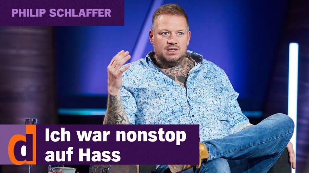 Philip Schlaffer ist tätowiert und trägt ein blau gemustertes Hemd und Jeans. Er sitzt während eines Interviews auf der Bühne. Der Text lautet "PHILIP SCHLAFFER" und "Ich war nonstop auf Hass", was auf einen einflussreichen Talkshow-Moment hindeutet.