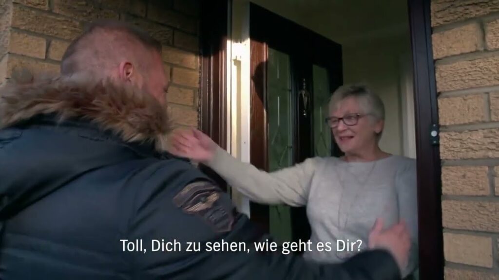 Ein Mann steht an einer Tür und wird von einer älteren Frau mit kurzen grauen Haaren und Brille begrüßt; sie lächelt und hebt den Arm zur Begrüßung. Deutsche Untertitel lauten: "Toll, Dich zu sehen, wie geht es Dir?"-Philip Schlaffer tritt herzlich ein.