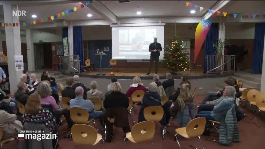 Eine Gruppe von Menschen sitzt auf Holzstühlen vor einer Bühne, auf der eine Person vor einer Projektionsfläche steht. Es gibt einen geschmückten Weihnachtsbaum und bunte Wimpel. Der Schauplatz scheint ein Gemeindesaal oder ein Veranstaltungsraum zu sein.